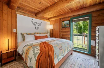 A cozy bedroom with warm wood walls, layered bedding, and a glass door looking out toward the deck and greenery.