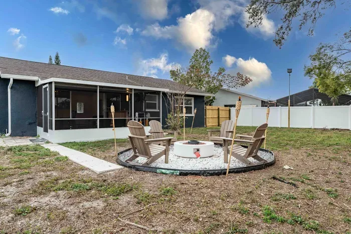A serene outdoor space anchored by a stylish fire pit, surrounded by comfy Adirondack chairs. The gravel circle creates a cozy gathering spot, perfect for evening storytelling or stargazing, all framed by lush greenery and a privacy fence.