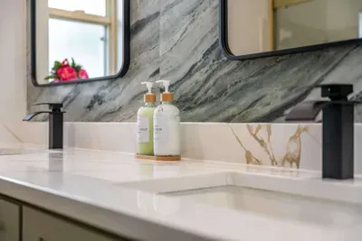 A stylish vanity with a sleek countertop and two modern mirrors reflects a minimalist design. Luxurious hand soaps sit on a wooden tray, adding a touch of sophistication against the elegant marble backdrop