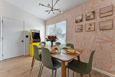 This photo presents the dining area from a cozy perspective, capturing the interplay between light and decor. The colorful tableware and floral arrangements create a cheerful vibe, while the vintage arcade game in the corner adds a playful twist.