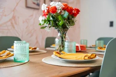 A beautifully arranged dining table set for a gathering, showcasing vibrant orange and white flowers in a stylish vase. The bright yellow napkins and cool blue glasses create a cheerful contrast against the wooden table, making the space feel warm,