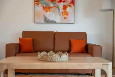 A cozy brown sofa adorned with vibrant orange cushions sits against a bright wall, offering a warm and inviting feel. Above the sofa, a modern abstract painting in shades of orange and cream adds a splash of creativity, while a simple wooden coffee.