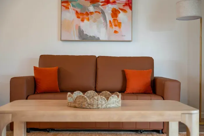 A cozy brown sofa adorned with vibrant orange cushions sits against a bright wall, offering a warm and inviting feel. Above the sofa, a modern abstract painting in shades of orange and cream adds a splash of creativity, while a simple wooden coffee.