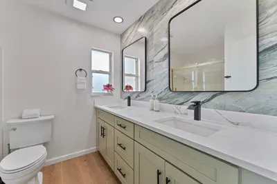 A spacious bathroom layout featuring dual sinks beneath a striking marble wall. Modern mirrors enhance the aesthetic, and natural light filters in through a window, adding warmth and brightness to this elegant retreat.