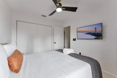 This bedroom showcases a fresh, airy ambiance with a beautifully made bed adorned with neatly arranged pillows. Vibrant orange curtains frame the window, while colorful wall art adds a playful touch, creating a delightful atmosphere for relaxation.