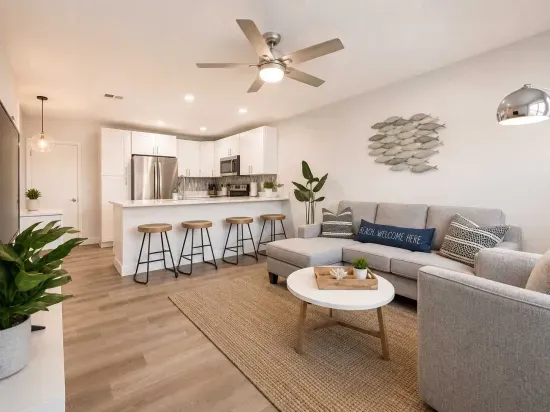 Bright and modern open-concept living space featuring a cozy sectional, sleek kitchen with bar seating, and stylish coastal décor. Natural light, warm wood floors, and soft neutrals create a welcoming retreat perfect for relaxing or entertaining.