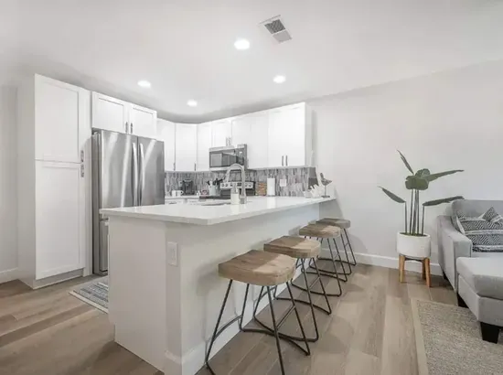 Sleek, contemporary kitchen with crisp white cabinetry, stainless steel appliances, and a spacious island with wooden bar stools. Bright lighting and modern finishes create an inviting space perfect for cooking, dining, and gathering with friends.