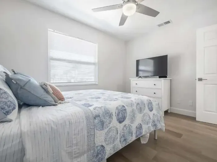 Cozy and bright bedroom featuring a comfortable bed with coastal-patterned bedding, a flat-screen TV, and calming neutral tones. The airy design and natural light create a peaceful retreat for rest and relaxation.