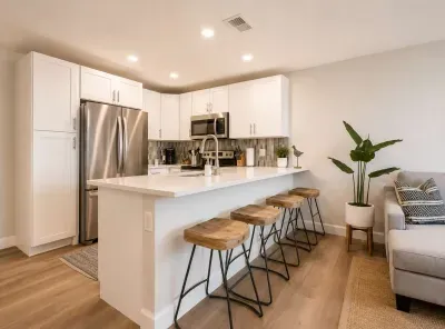 Sleek, contemporary kitchen with crisp white cabinetry, stainless steel appliances, and a spacious island with wooden bar stools. Bright lighting and modern finishes create an inviting space perfect for cooking, dining, and gathering with friends.