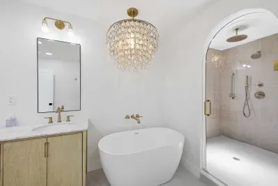 Modern spa-style bathroom with a freestanding tub, arched glass shower, and warm wood vanity. Gold fixtures and sleek finishes create a luxurious, calming retreat designed for pure relaxation and elegant coastal comfort.