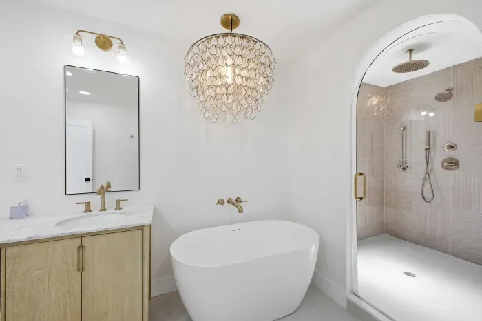 Modern spa-style bathroom with a freestanding tub, arched glass shower, and warm wood vanity. Gold fixtures and sleek finishes create a luxurious, calming retreat designed for pure relaxation and elegant coastal comfort.