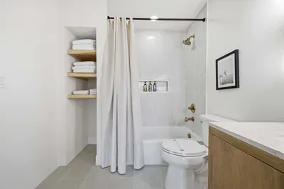 Bright, modern bathroom featuring a tiled shower-tub combo, open shelving, and a sleek wood vanity. Simple yet elegant, this coastal-inspired space offers comfort and style for a refreshing start or end to your day.