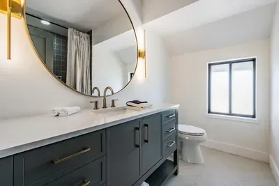Sophisticated bathroom featuring a bold navy vanity, gold fixtures, and a stunning round mirror. Bright and airy with modern coastal charm—this stylish space offers a refreshing, spa-like experience.