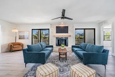 Bright and elegant living room featuring plush blue sofas, a cozy fireplace, and large windows framing palm-filled views. Perfect for gathering or unwinding, this stylish space blends modern design with coastal charm and natural light.