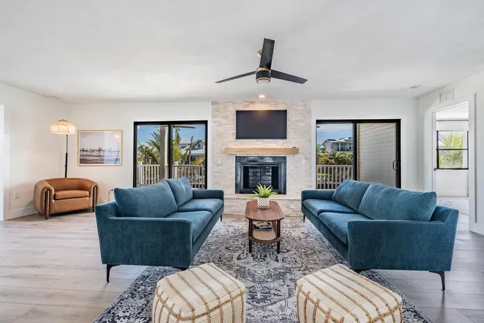 Bright and elegant living room featuring plush blue sofas, a cozy fireplace, and large windows framing palm-filled views. Perfect for gathering or unwinding, this stylish space blends modern design with coastal charm and natural light.