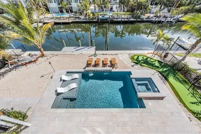 Gorgeous waterfront pool area featuring a sparkling pool, spa, and sun loungers overlooking the canal. Surrounded by palm trees and a putting green, this tropical retreat is perfect for relaxation and outdoor fun.