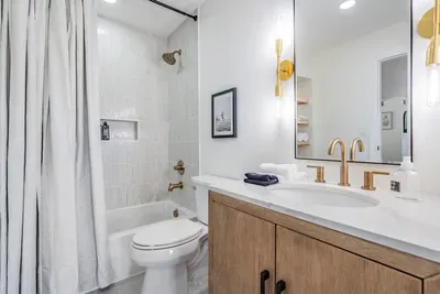 Modern bathroom with sleek gold fixtures, a light wood vanity, and a tiled shower-tub combo. Bright and stylish, this space combines coastal elegance with everyday comfort for a refreshing retreat.
