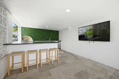 Modern outdoor lounge with full bar, sleek countertops, and seating for guests. Enjoy the wall-mounted TV, stylish green accent wall, and open-air design—perfect for entertaining, watching the game, or relaxing poolside in comfort.