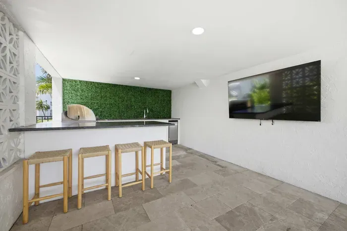 Modern outdoor lounge with full bar, sleek countertops, and seating for guests. Enjoy the wall-mounted TV, stylish green accent wall, and open-air design—perfect for entertaining, watching the game, or relaxing poolside in comfort.