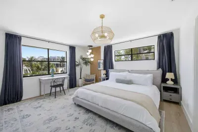 Elegant primary bedroom with a king bed, chic lighting, and soothing coastal tones. Large windows fill the space with natural light and views of palm trees, creating a tranquil retreat perfect for rest and relaxation.