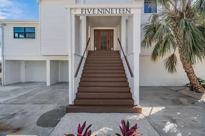Elegant entryway framed by tropical palms and modern architecture. The grand staircase leads to double doors, welcoming guests into a bright coastal retreat that exudes style, comfort, and a touch of island sophistication.