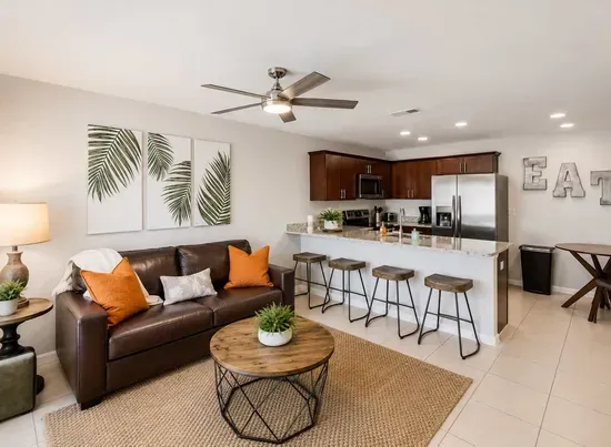 Stylish open-concept living area featuring a cozy leather sofa, chic décor, and a modern kitchen with stainless-steel appliances. Enjoy casual dining at the breakfast bar or unwind under the ceiling fan in this bright, inviting space.