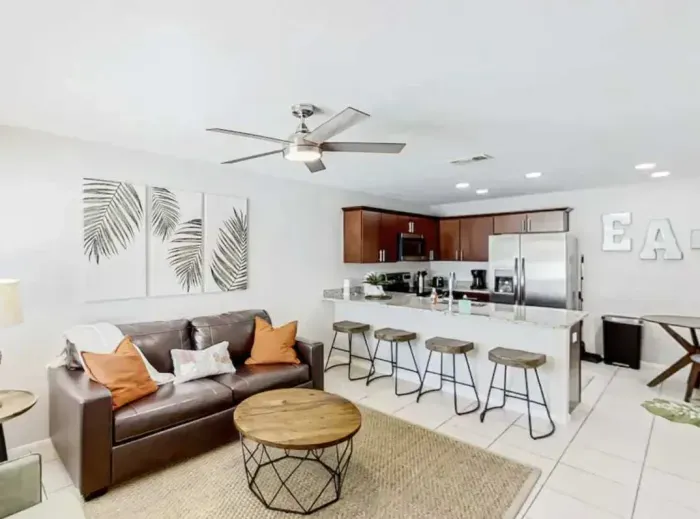 Stylish open-concept living area featuring a cozy leather sofa, chic décor, and a modern kitchen with stainless-steel appliances. Enjoy casual dining at the breakfast bar or unwind under the ceiling fan in this bright, inviting space.