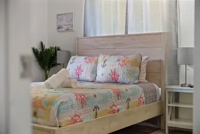 Relax in this cozy queen bedroom featuring fresh linens, coastal-inspired bedding, and soft natural light. Thoughtful touches like bedside lighting and clean, modern furnishings create a comfortable retreat—perfect for restful nights after pool days