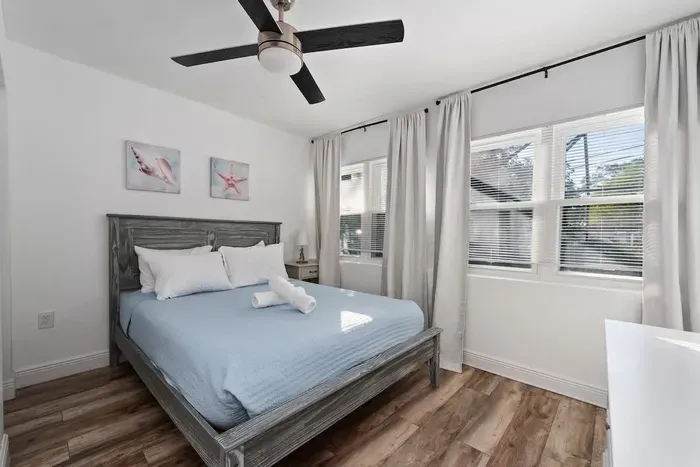 Bright and relaxing queen bedroom with warm wood floors, soft natural light, and a ceiling fan for added comfort. Enjoy fresh linens, modern décor, and a peaceful vibe—perfect for a restful night’s sleep after pool time and sunny Florida days.