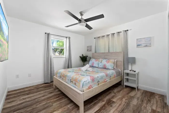 Charming and bright queen bedroom with coastal-style décor, warm wood floors, and a ceiling fan for added comfort. Enjoy fresh linens, soft lighting, and a peaceful layout—perfect for unwinding and recharging after a fun day in the sun.