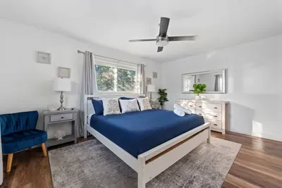 Bright and serene king bedroom with stylish décor, warm wood floors, and plenty of natural light. Enjoy a comfortable bed with fresh linens, a ceiling fan for added comfort, and a spacious layout—perfect for relaxing and recharging in total comfort.