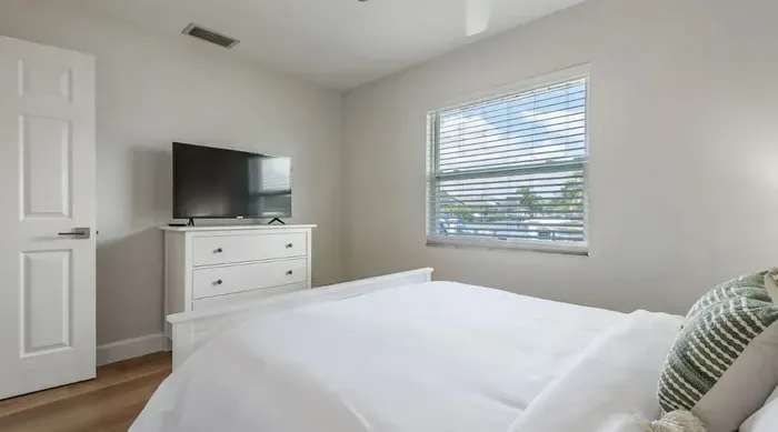 Bright and cozy bedroom with a plush bed, flat-screen TV, and large window letting in natural light. Simple, elegant design for a peaceful and comfortable stay.