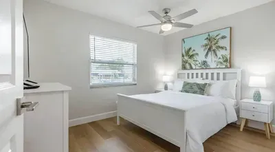 Coastal-inspired bedroom with a comfy white bed, tropical artwork, and soft natural light. Calming tones and modern touches create a relaxing retreat perfect for unwinding after a sunny day.