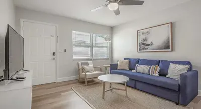 Inviting living area with a chic blue sofa, cozy armchair, and natural textures throughout. Bright windows fill the space with sunlight, creating a relaxing coastal vibe perfect for unwinding or watching TV in comfort.