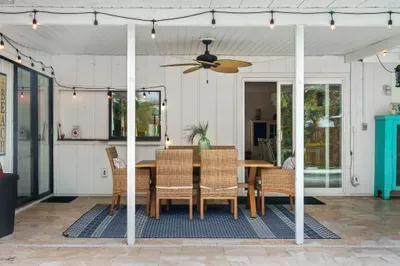 This image features an elegant outdoor dining area with a wooden table surrounded by woven chairs, ideal for al fresco meals. A large ceiling fan hangs overhead, and string lights add a warm ambiance