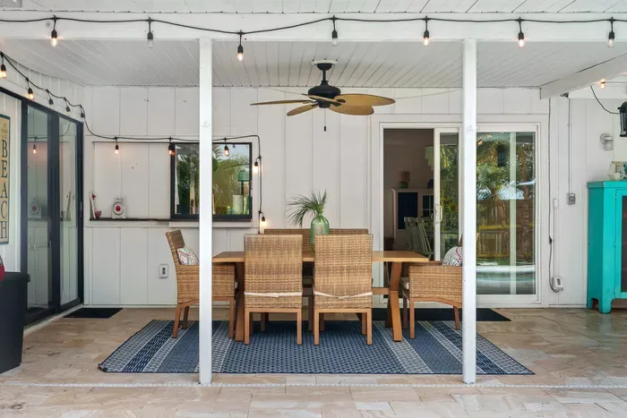 This image features an elegant outdoor dining area with a wooden table surrounded by woven chairs, ideal for al fresco meals. A large ceiling fan hangs overhead, and string lights add a warm ambiance