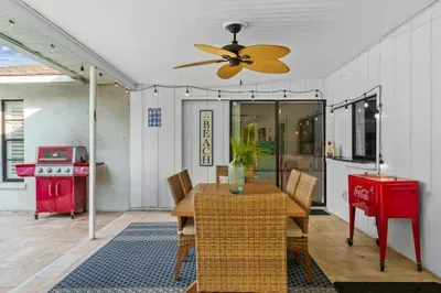 Charming outdoor dining area, highlighted by a large wooden table surrounded by wicker chairs. A vibrant green potted plant adds a fresh touch to the table. The space is well-lit with string lights above and a ceiling fan, enhancing the relaxed vibe.