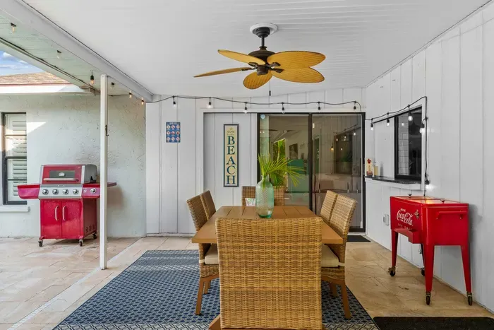 Charming outdoor dining area, highlighted by a large wooden table surrounded by wicker chairs. A vibrant green potted plant adds a fresh touch to the table. The space is well-lit with string lights above and a ceiling fan, enhancing the relaxed vibe.