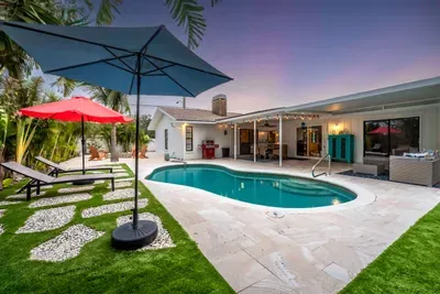 This angle offers a broader view of the pool and surrounding patio, showcasing a modern outdoor living space. The red and blue umbrellas shade the loungers, and the lush greenery enhances the tropical ambiance.