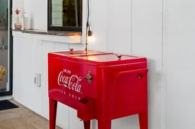 Coca-Cola cooler on legs, adding a fun nostalgic touch to the outdoor setting. The cooler gleams under a hanging bulb, inviting guests to enjoy drinks while relaxing.