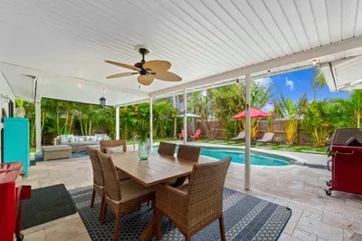 Beautiful outdoor patio with a clear view of a sparkling pool surrounded by lush greenery. A large wooden dining table with wicker chairs creates a welcoming dining space, while a cozy seating area is visible in the background
