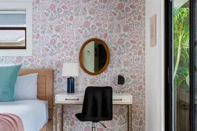 This photo captures a cozy bedroom corner with a floral wallpaper accent wall. A wooden bed frame is paired with soft blue and pink cushions, while a simple black chair sits at a small desk beneath a round mirror
