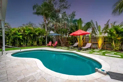 This photo highlights two loungers beneath a bright red umbrella, framed by tall palm trees and lush foliage. The clean lines of the stone pathway lead to the pool, while the bright blue sky creates a cheerful atmosphere, inviting guests to unwind