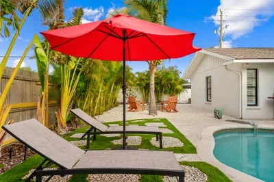 This image captures a beautifully landscaped pool area with a clear blue swimming pool at the center. Two red umbrellas provide shade over black loungers, while pink accents and vibrant plants surround the area, creating a tropical oasis.