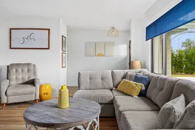The image captures a cheerful living room featuring a soft beige sectional sofa, complemented by a sunny yellow vase as a centerpiece. A cozy armchair sits nearby, and the bright blue window covering allows natural light to flow in.