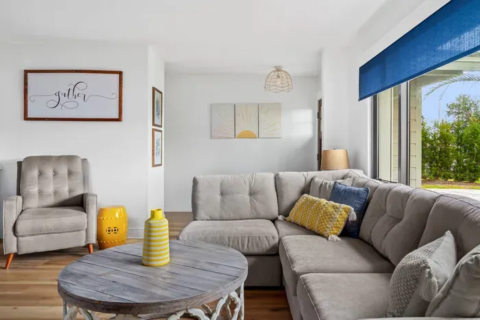 The image captures a cheerful living room featuring a soft beige sectional sofa, complemented by a sunny yellow vase as a centerpiece. A cozy armchair sits nearby, and the bright blue window covering allows natural light to flow in.