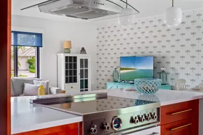 This photo offers a glimpse of the kitchen from a different angle, highlighting a stainless steel gas range and a mounted hood. A vibrant television displays a serene beach view, adding to the light, airy feel.