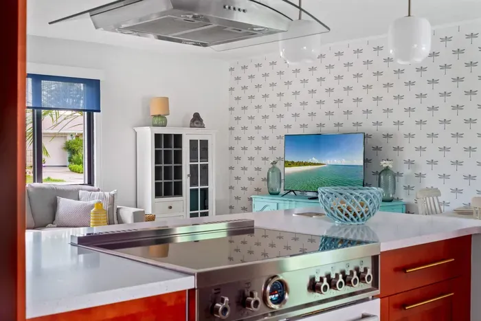 This photo offers a glimpse of the kitchen from a different angle, highlighting a stainless steel gas range and a mounted hood. A vibrant television displays a serene beach view, adding to the light, airy feel.