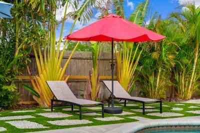 Featuring the same loungers and red umbrella, this angle highlights the lush tropical backdrop and a cozy atmosphere. The combination of grass and pebbled pathways adds an inviting texture to the outdoor space.