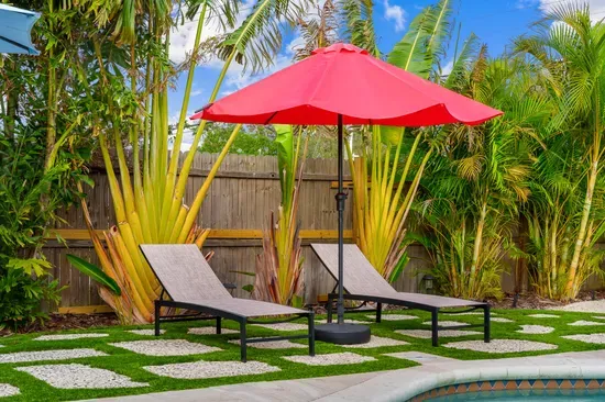 Featuring the same loungers and red umbrella, this angle highlights the lush tropical backdrop and a cozy atmosphere. The combination of grass and pebbled pathways adds an inviting texture to the outdoor space.